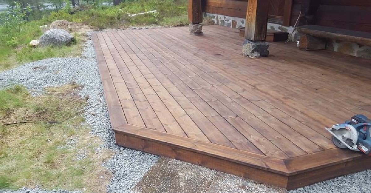 Nybygd terrasse i beiset tre med grus rundt, festet til en hytte med tømmerstolper.