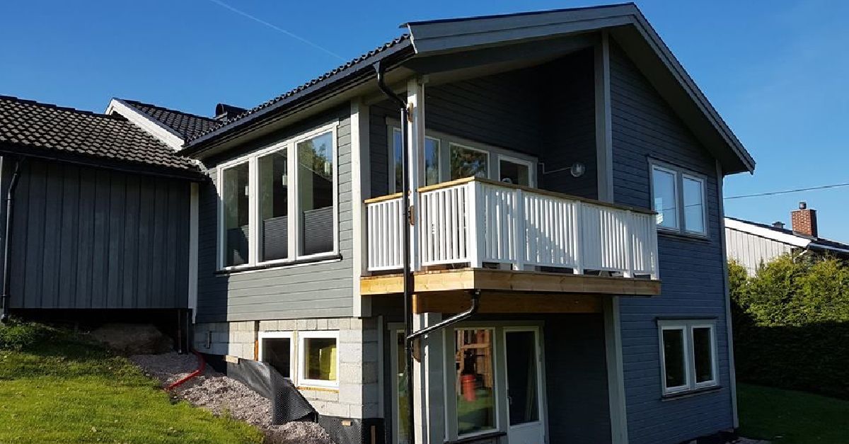 Moderne enebolig med mørkegrå kledning, hvite vinduer og en liten balkong. Hagen har grønt gress og grus langs grunnmuren.