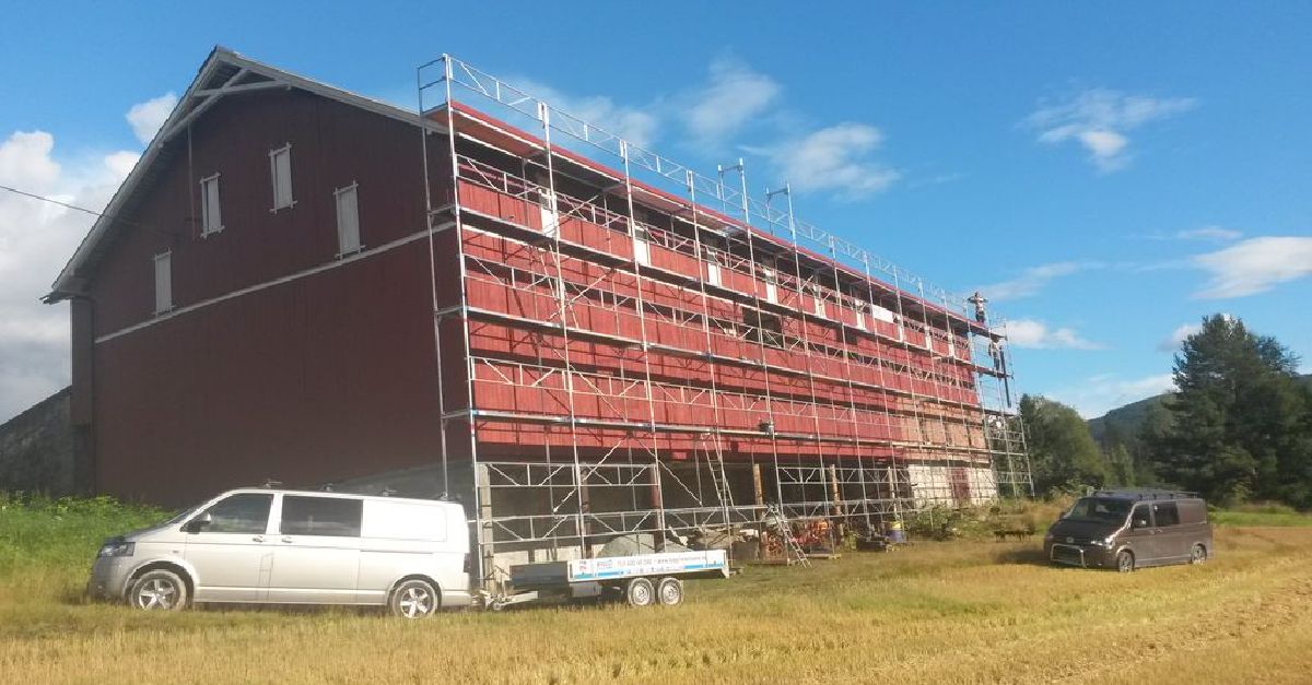 Rød låve med stillas langs veggen, omgitt av biler og utstyr på en åker.